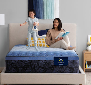 Mother reading a book while a child jumps happily on a Serta Classic Latex mattress with blue top and navy floral-pattern border on a beige upholstered bed frame