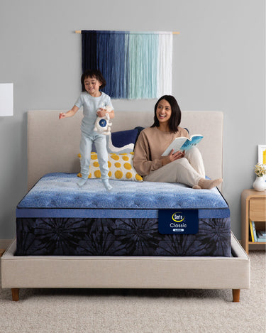 Mother reading a book while a child jumps happily on a Serta Classic Latex mattress with blue top and navy floral-pattern border on a beige upholstered bed frame