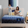 Mother reading a book while a child jumps happily on a Serta Classic Latex mattress with blue top and navy floral-pattern border on a beige upholstered bed frame