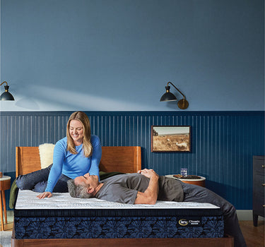 Couple relaxing on a Serta Classic Luxury mattress with white tufted top and navy floral-pattern border on a wooden bed frame in a blue-walled bedroom