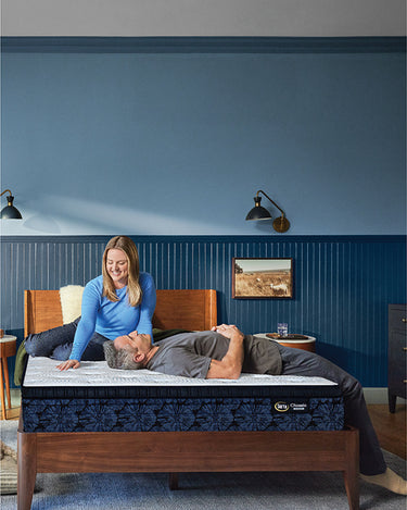 Couple relaxing on a Serta Classic Luxury mattress with white tufted top and navy floral-pattern border on a wooden bed frame in a blue-walled bedroom