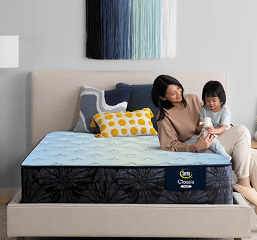 Mother and child sitting together on a Serta Classic Plus mattress with light blue quilted top on a beige upholstered bed frame in a cozy bedroom