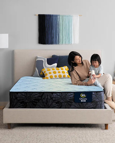 Mother and child sitting together on a Serta Classic Plus mattress with light blue quilted top on a beige upholstered bed frame in a cozy bedroom