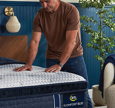 Man pressing down on the pillow-top surface of a Serta iComfort Eco mattress on a wooden bed frame in a blue-accented bedroom setting