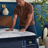 Man pressing down on the pillow-top surface of a Serta iComfort Eco mattress on a wooden bed frame in a blue-accented bedroom setting