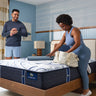 Couple in a blue-toned bedroom with a woman sitting on a Serta iComfort mattress featuring white and navy swirl-patterned fabric on a wooden bed frame