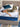 A woman relaxing on a Serta iLatex mattress with a white tufted top and navy blue quilted border, placed on a grey upholstered bed frame