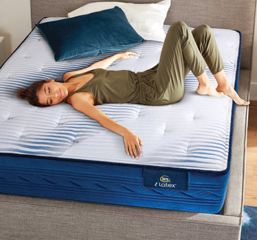 A woman relaxing on a Serta iLatex mattress with a white tufted top and navy blue quilted border, placed on a grey upholstered bed frame