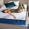 A woman relaxing on a Serta iLatex mattress with a white tufted top and navy blue quilted border, placed on a grey upholstered bed frame