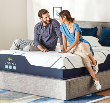 Smiling couple sitting together on a Serta iSeries mattress with white diagonal-striped top and navy blue base on a grey upholstered bed frame in a modern bedroom