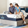 Smiling couple sitting together on a Serta iSeries mattress with white diagonal-striped top and navy blue base on a grey upholstered bed frame in a modern bedroom