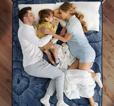 Overhead view of a family — parents and young child — sleeping together on a Serta Perfect Sleeper Hybrid mattress with navy blue floral radial-pattern fabric on a wooden bed frame