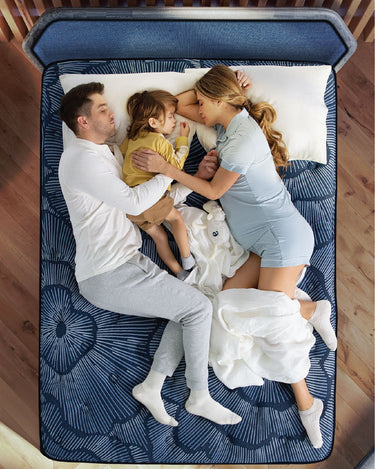 Overhead view of a family — parents and young child — sleeping together on a Serta Perfect Sleeper Hybrid mattress with navy blue floral radial-pattern fabric on a wooden bed frame