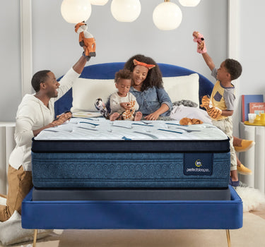 Happy family with two children playing on a Serta Perfect Sleeper mattress with white dash-quilted top and blue oval-patterned border on a navy upholstered bed frame